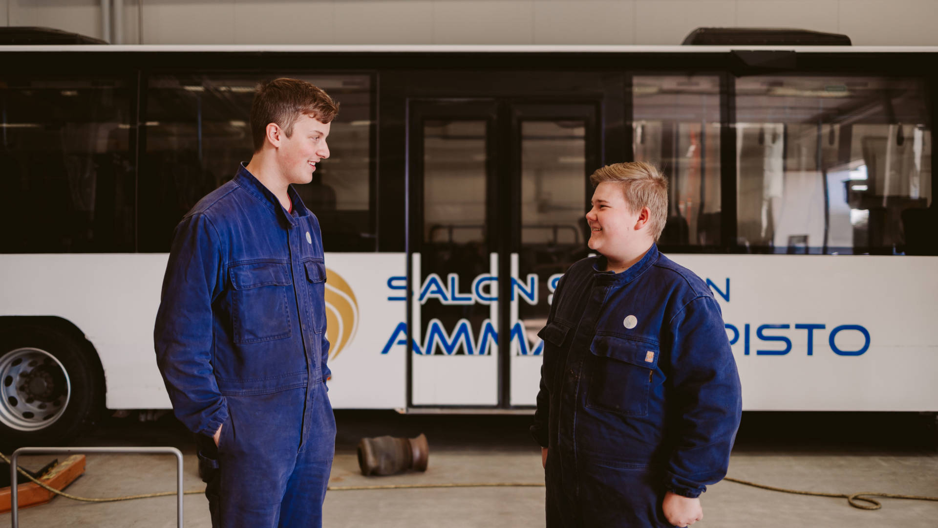 Kaksi nuorta autoalan opiskelijaa keskustelevat työsalissa korjatun linja-auton edessä hymyillen.