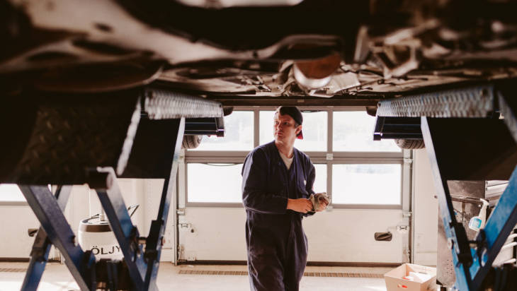 Autoalan nuori opiskelija pohtii ilmaan nostetun auton takana auton vikoja valoisassa korjaamossa.