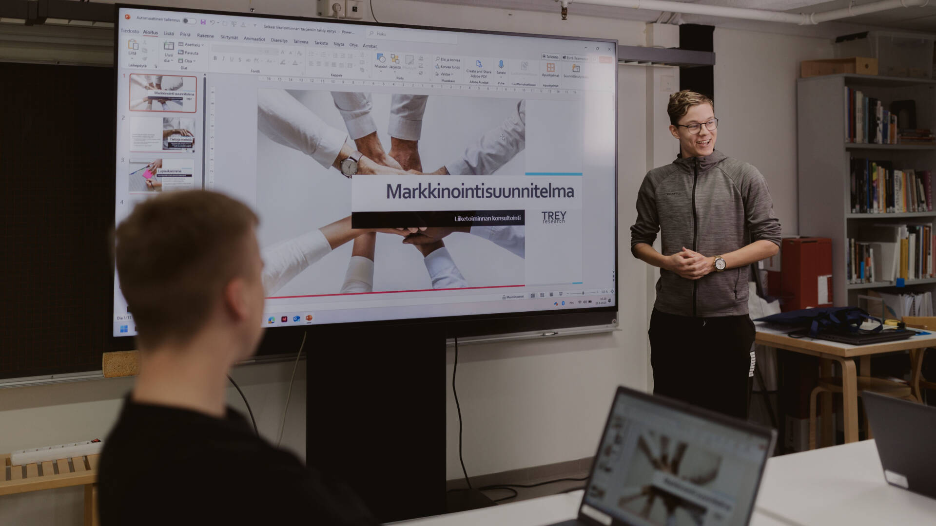 Liiketoiminnan alan opiskelija esittelee markkinointisuunnitelmaa luokan edessä muiden opiskelijoiden kuunnellessa.