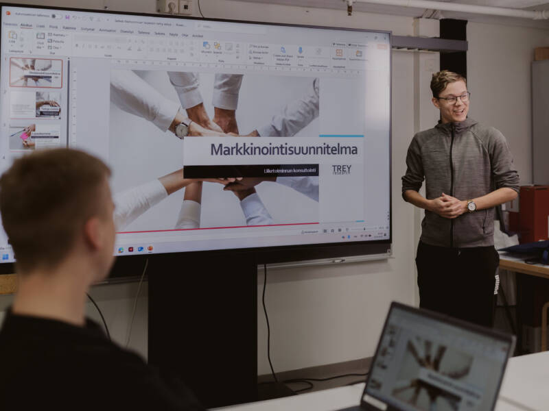 Liiketoiminnan alan opiskelija esittelee markkinointisuunnitelmaa luokan edessä muiden opiskelijoiden kuunnellessa.