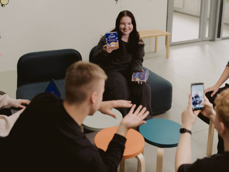 Liiketoiminnan alan opiskelija ottaa valokuvaa toisesta opiskelijasta, joka esittelee tuotepakkauksia oppimisympäristössä.