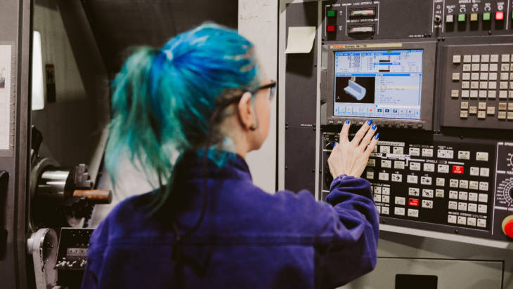 Metallialan opiskelija sinisissä työhaalareissa tarkistaa seinän näytöltä tietoja laitteesta.