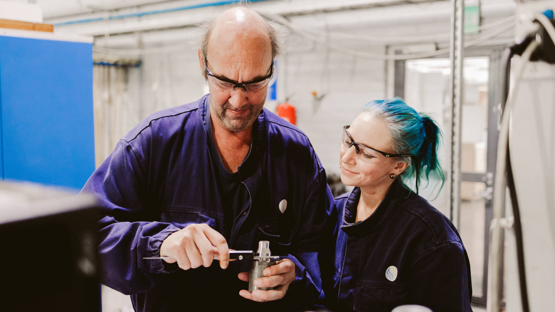 Metallialan opettaja näyttää nuoremmalle oppilaalle esimerkkiä työkalun käytöstä ja oppilas seuraa turvalasien kanssa vierestä.