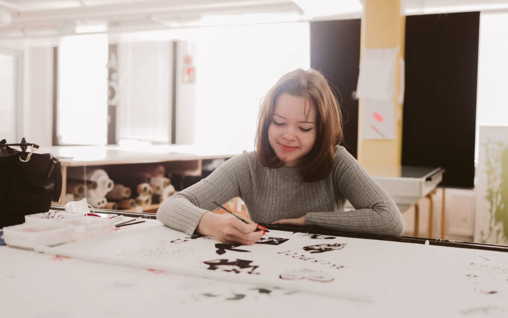 Nuori ja iloinen tekstiilialan opiskelija suunnittelee kankaaseen painettavia kuvia paperille.