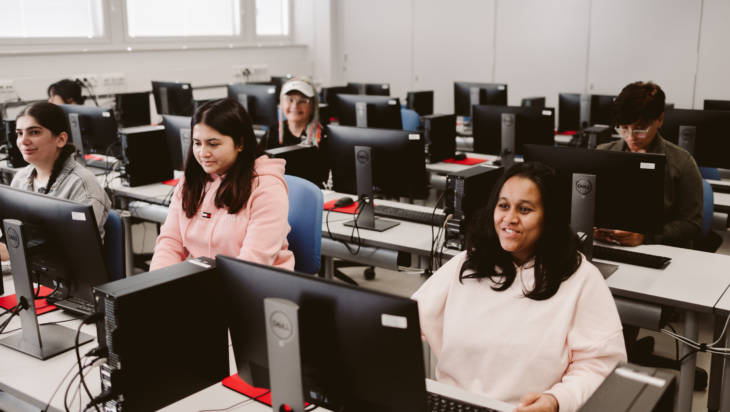 Joukko iloisia kansainvälisiä opiskelijoita istuu tietokoneiden takana isossa luokkatilassa.