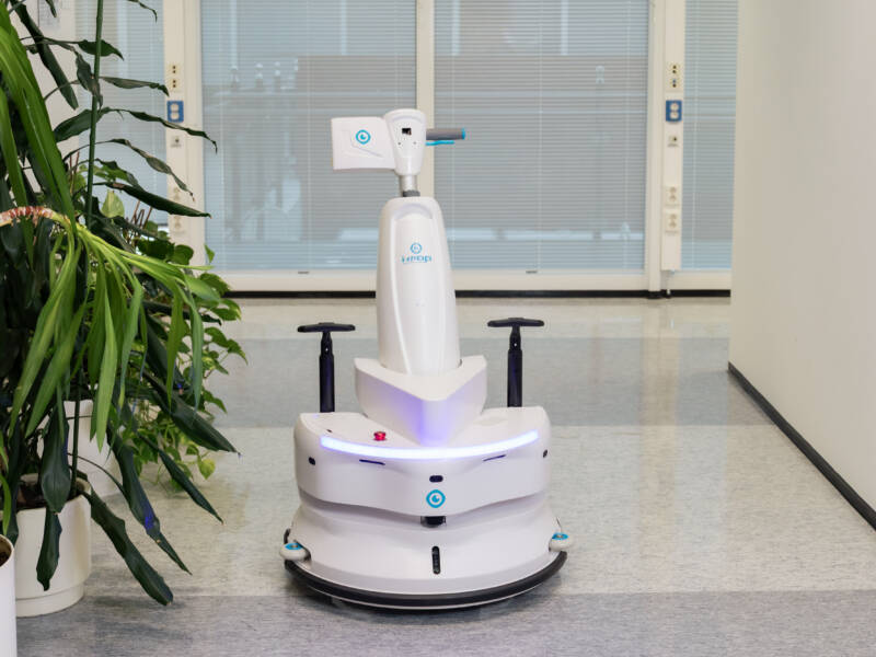 Robottiohjattu lattianpesurobotti oppilaitoksen käytävässä viherkasvien vierellä.