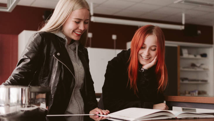 Kaksi nuorta opiskelijatyttöä tutkii yhdessä pöydän ääressä kirjaa.